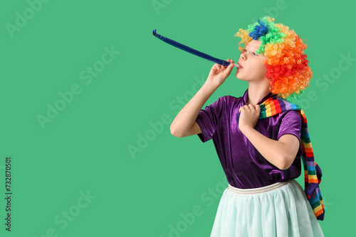 Funny girl in clown costume with party whistle on green background. April Fool's Day celebration