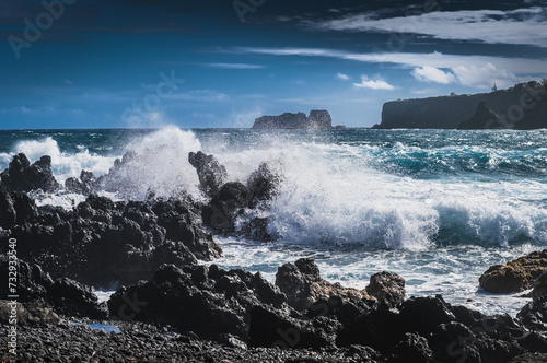 View of waves crushing over black rocks; distant shore in background