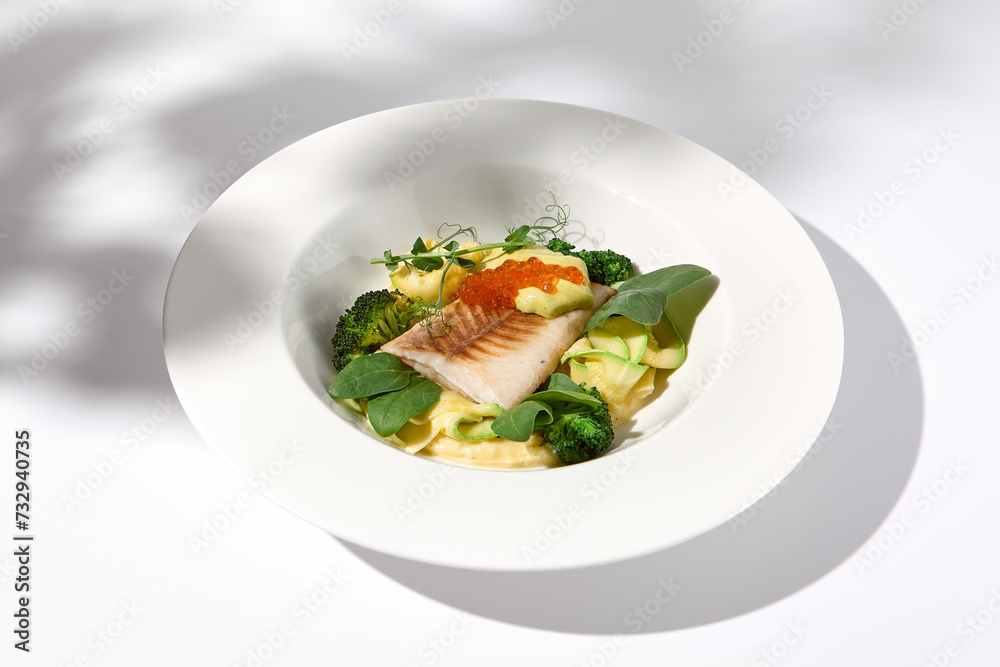 Cod fillet with caviar and zucchini-broccoli garnish, side view on white plate with delicate leaf shadow Cod fillet with caviar and zucchini-broccoli garnish, side view on white plate with delicate leaf shadow