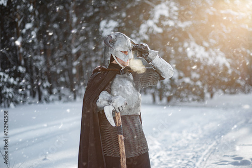 Portrait of a medieval fantasy warrior in a horned helmet, steel breastplate, chain mail with a two-handed ax in his hands, posing against the backdrop of sunset in a winter forest.