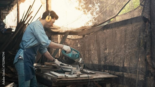Handsome young craftsman wearing safety glasses and hearing protection headphones is using a wood saw to cut pieces, Carpenter or furniture designer is working in a workshop.	 photo