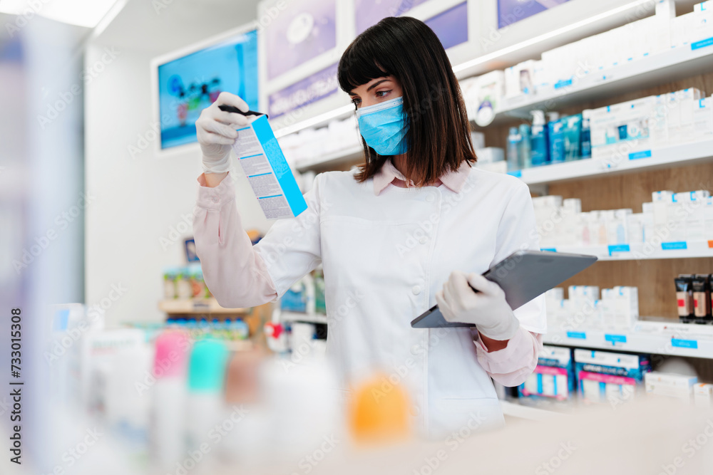 Beautiful pharmacist with protective face mask working and standing in a drug store and doing a stock take. Portrait of a positive healthcare worker or a chemist at his work. Beautiful pharmacist with protective face mask working and standing in a drug store and doing a stock take. Portrait of a positive healthcare worker or a chemist at his work.