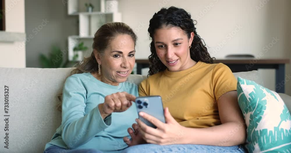 Young 25s daughter showing new mobile application to mature 55s mother sit on sofa staring at cellphone screen watching funny videos, laughing enjoy family leisure on internet. Modern tech, new apps Young 25s daughter showing new mobile application to mature 55s mother sit on sofa staring at cellphone screen watching funny videos, laughing enjoy family leisure on internet. Modern tech, new apps