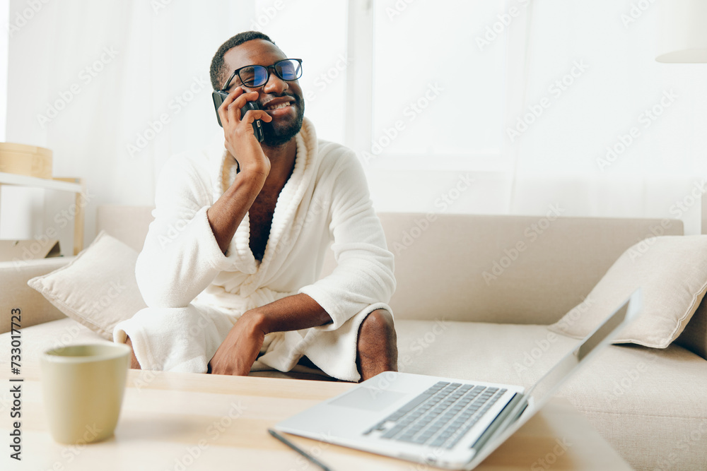 Smiling African American Man Working on Laptop in Cozy Home Office, Talking on Phone, Wearing Bathrobe, Surrounded by modern technology and wireless devices Smiling African American Man Working on Laptop in Cozy Home Office, Talking on Phone, Wearing Bathrobe, Surrounded by modern technology and wireless devices