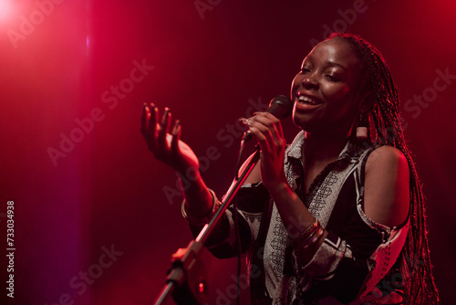Waist up portrait of talented Black woman singing into microphone performing on stage in dim red lights copy space