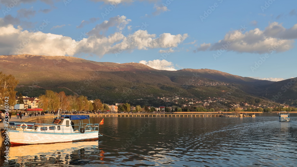 Tourboat moored on Kej Makedonija promenade close to a long pier jutting into the lake reaching an old swimming court ruins. Ohrid-North Macedonia-333