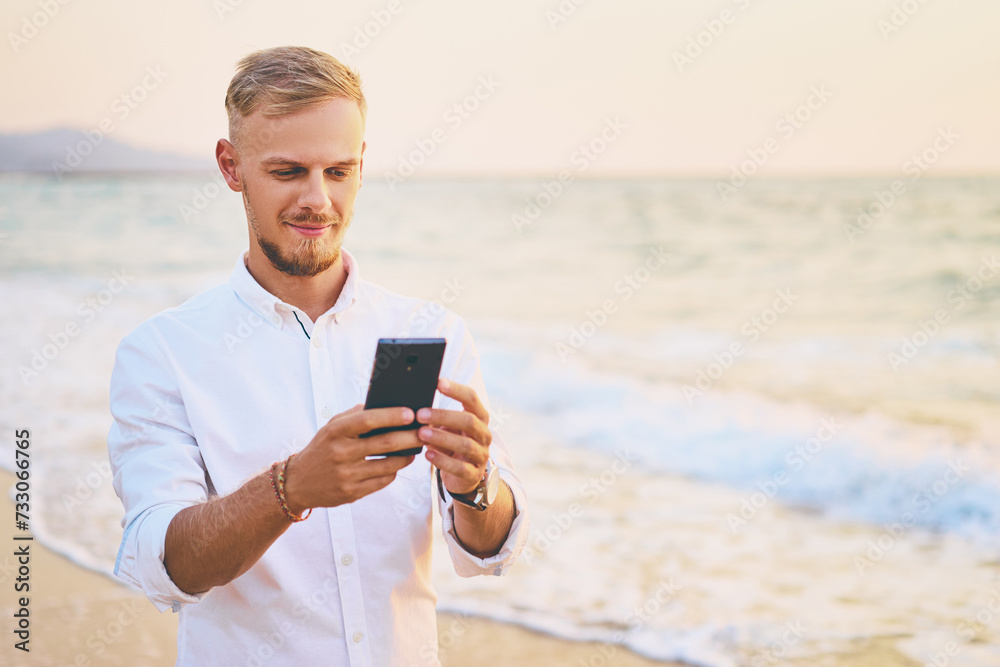 Vacation and technology. Portrait of young handsome man using smartphone on the sea shore.