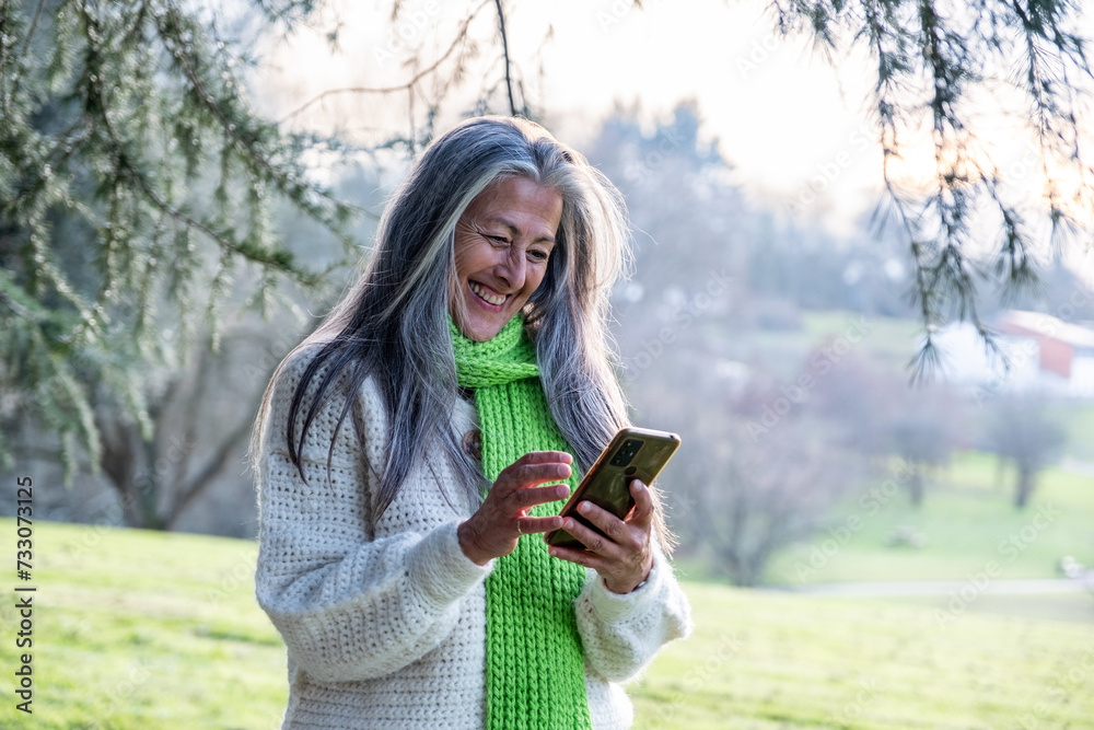 A modern and attractive middle-aged woman with long white hair looking at her mobile phone in a park on a wintry day. A modern and attractive middle-aged woman with long white hair looking at her mobile phone in a park on a wintry day.