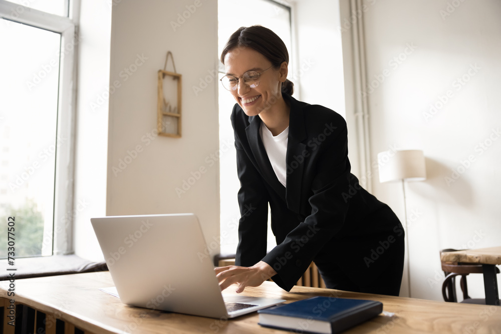 Positive ambitious young business woman working at laptop, standing at table with computer, typing, smiling, laughing, enjoying online communication, successful job connection on Internet Positive ambitious young business woman working at laptop, standing at table with computer, typing, smiling, laughing, enjoying online communication, successful job connection on Internet