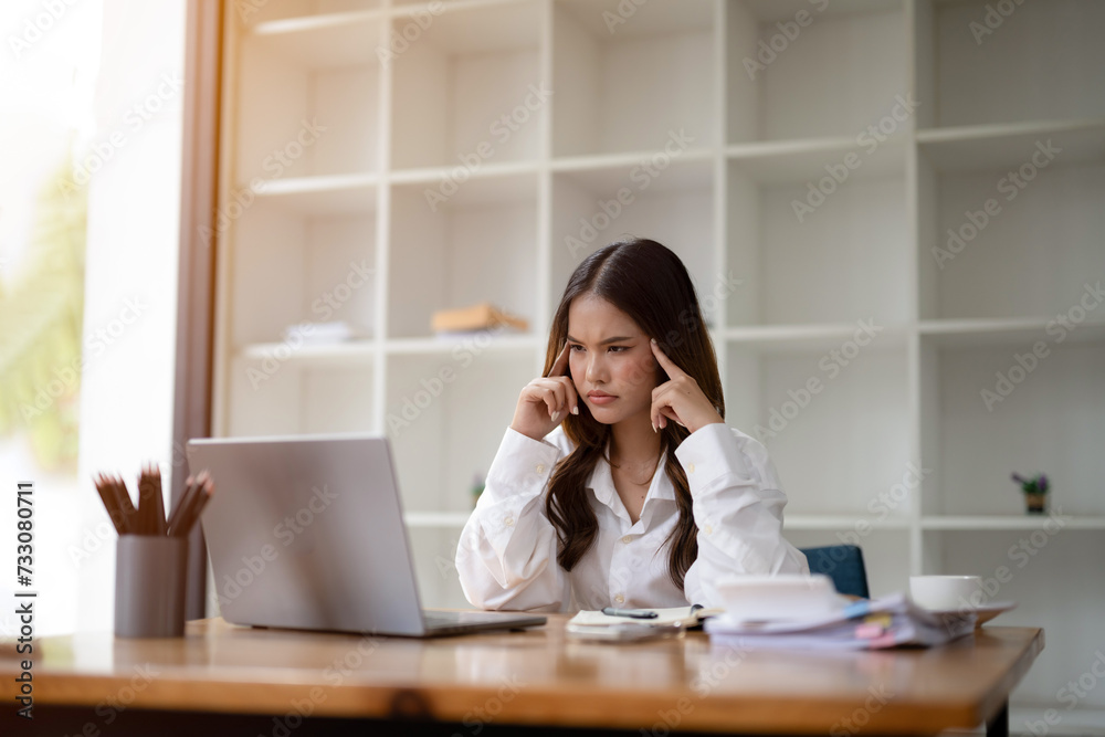 © Wasana - Focused businesswoman feeling a headache, massaging temples while working on a computer in a modern office..