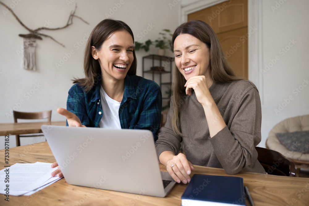 Two happy young businesswomen discussing creative strategy for project at laptop, sitting at computer, smiling, laughing, watching online Internet content, brainstorming on business ideas Two happy young businesswomen discussing creative strategy for project at laptop, sitting at computer, smiling, laughing, watching online Internet content, brainstorming on business ideas