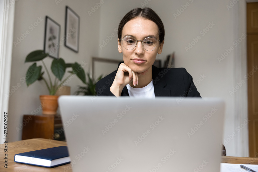 Positive focused thoughtful young business woman using online communication for job, working at laptop in home office, touching chin, looking at computer through stylish glasses, thinking Positive focused thoughtful young business woman using online communication for job, working at laptop in home office, touching chin, looking at computer through stylish glasses, thinking