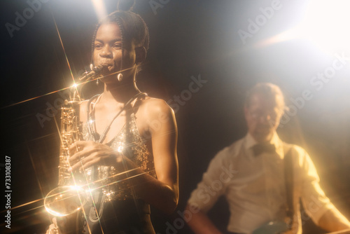 Portrait of Black young woman playing saxophone with jazz band on stage in hazy nightclub with golden lens flare accents copy space