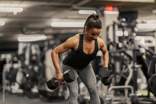 A strong sportswoman is lifting weights and bending while doing strength workouts in a gym.