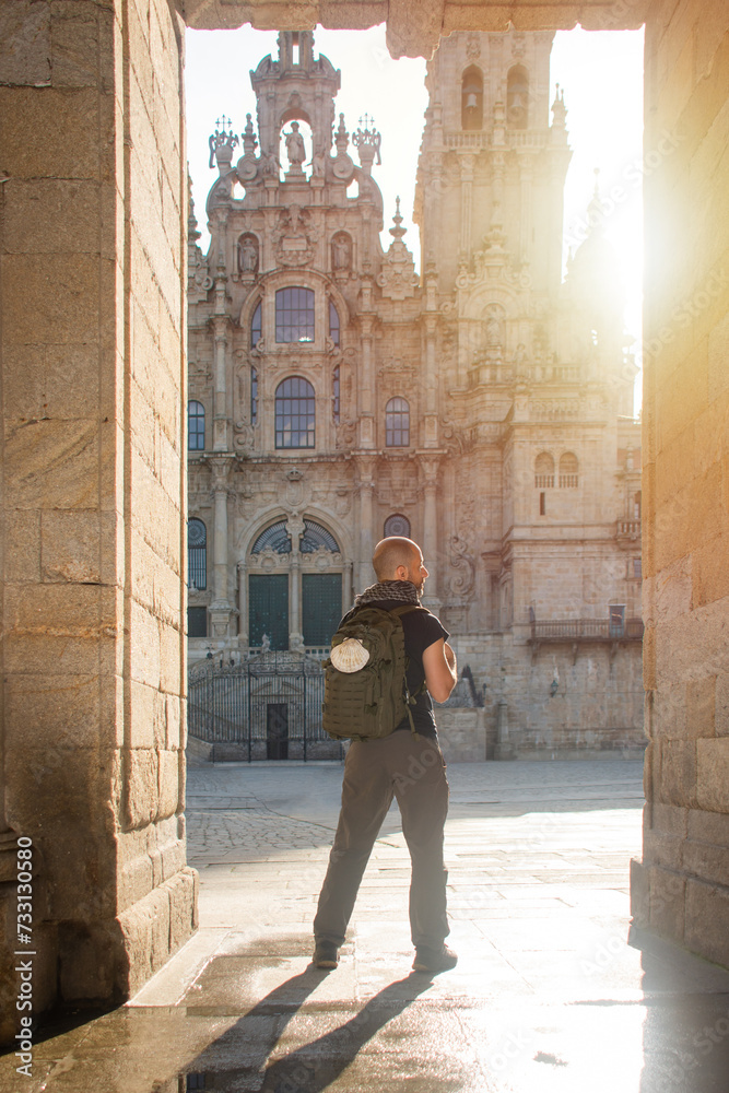 A mancarries a backpack adorned with a scallop shell, a. He is found framed in a stone door, entering the square of the Cathedral of Santiago de Compostela
