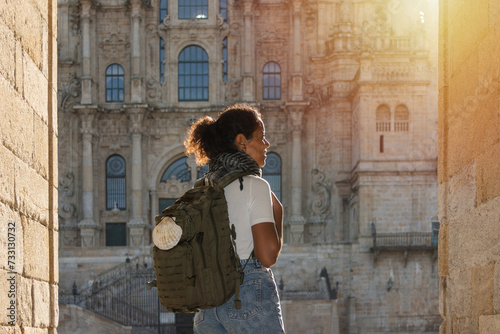 An African pilgrim woman, seen from behind, carrying a backpack adorned with a scallop shell symbol, entering the square of Santiago Cathedral illuminated by the morning sun
