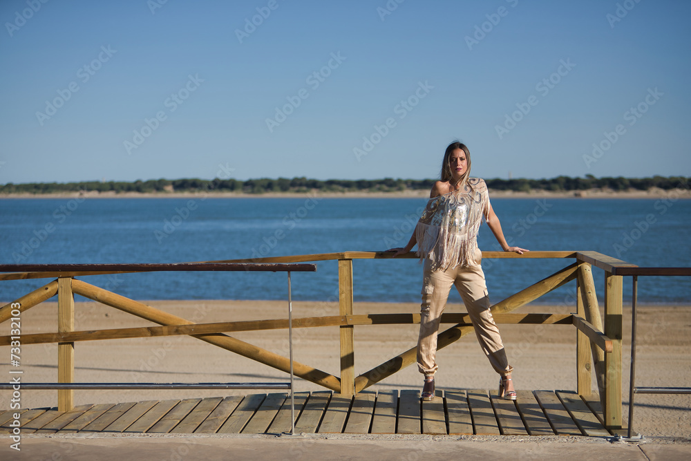 Young and beautiful woman with sequined and fringed shirt, leaning on a wooden railing, posing looking at the camera with the sea in the background. Concept beauty, fashion, trend, empowerment.