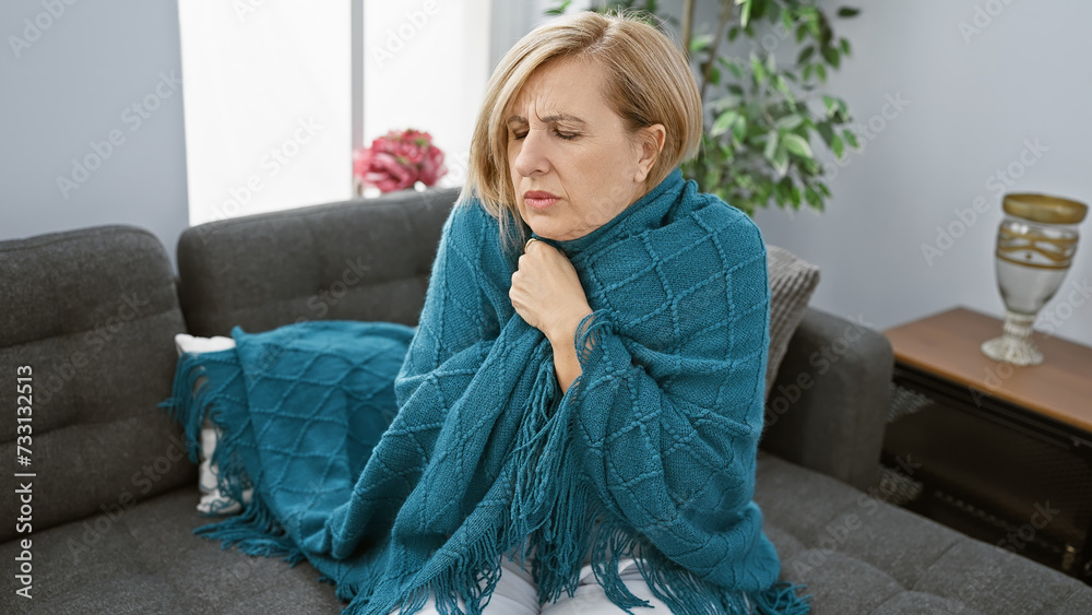 Naklejka premium Mature caucasian woman feeling unwell wrapped in a blanket sitting on her living room couch indoors.