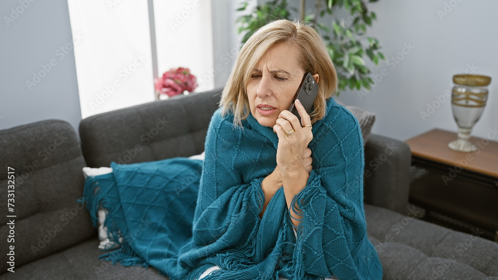A concerned middle-aged woman talks on a smartphone in a cozy living room, wrapped in a blue blanket. A concerned middle-aged woman talks on a smartphone in a cozy living room, wrapped in a blue blanket.