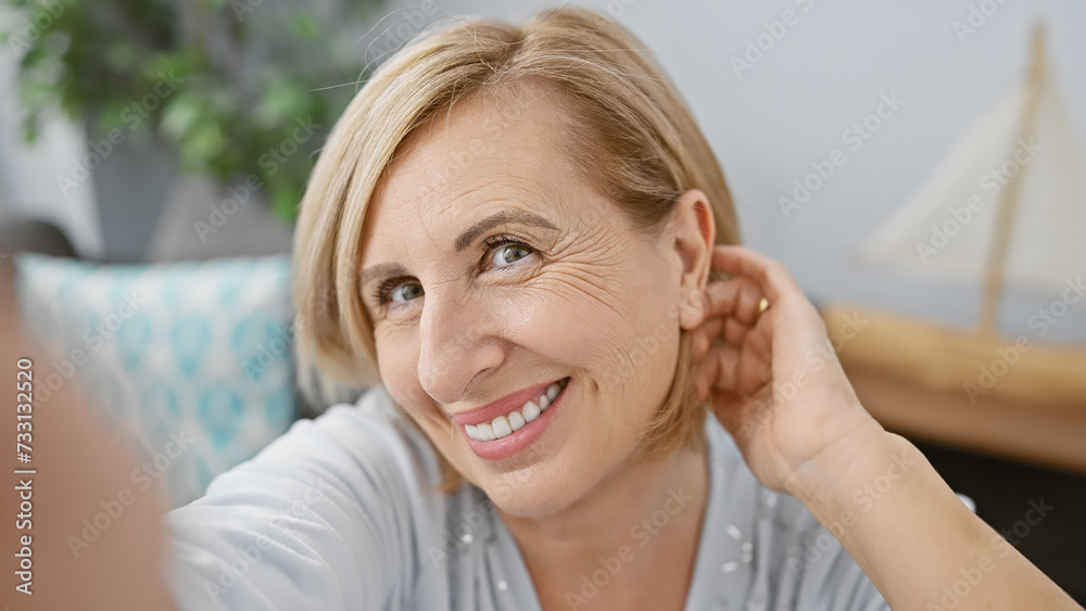 Fototapeta premium Middle-aged caucasian woman smiles warmly in a cozy living room interior, exuding casual elegance and comfort.