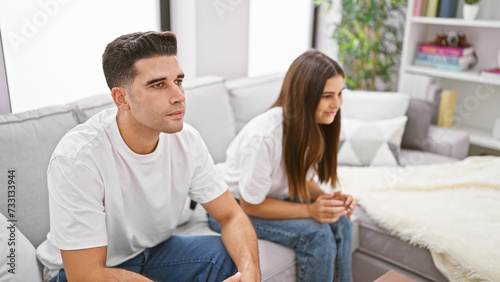 A contemplative couple sits apart on a sofa in their modern living room, depicting potential relationship issues.