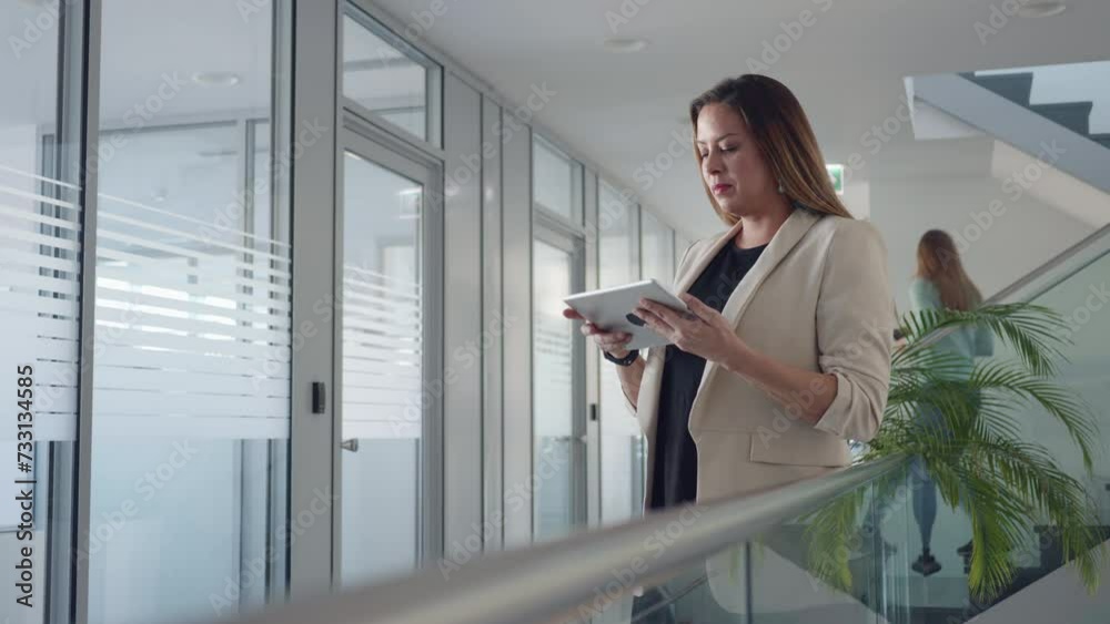 Female company worker tapping on digital tablet while standing in office corridor. Attractive businesswoman using portable gadget at workplace hall, handheld medium shot