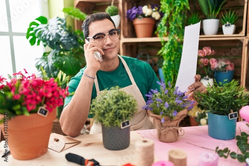Young hispanic man florist talking on smartphone reading document at flower shop