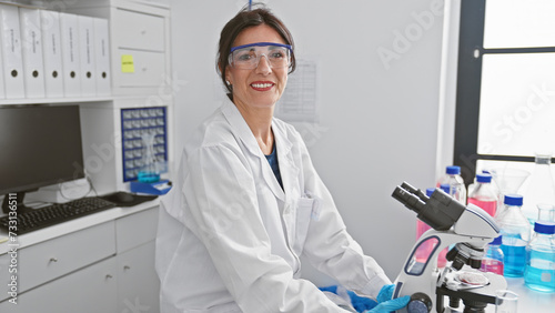 A smiling woman in a lab coat and protective glasses working with a microscope in a modern laboratory setting.