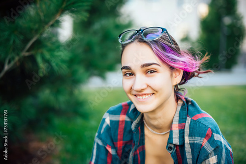 A beautiful smiling girl with glasses, using a smartphone, sitting outside in the park on the green grass. A pretty young girl is chatting with friends on a mobile phone