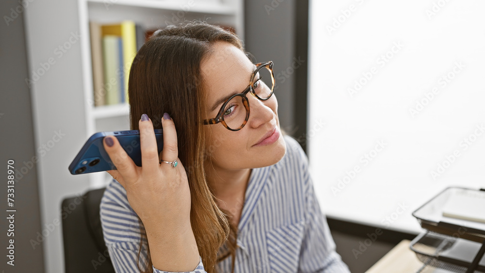 A young woman with brunette hair and glasses on a phone in a well-lit office interior. A young woman with brunette hair and glasses on a phone in a well-lit office interior.