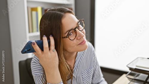 A young woman with brunette hair and glasses on a phone in a well-lit office interior.