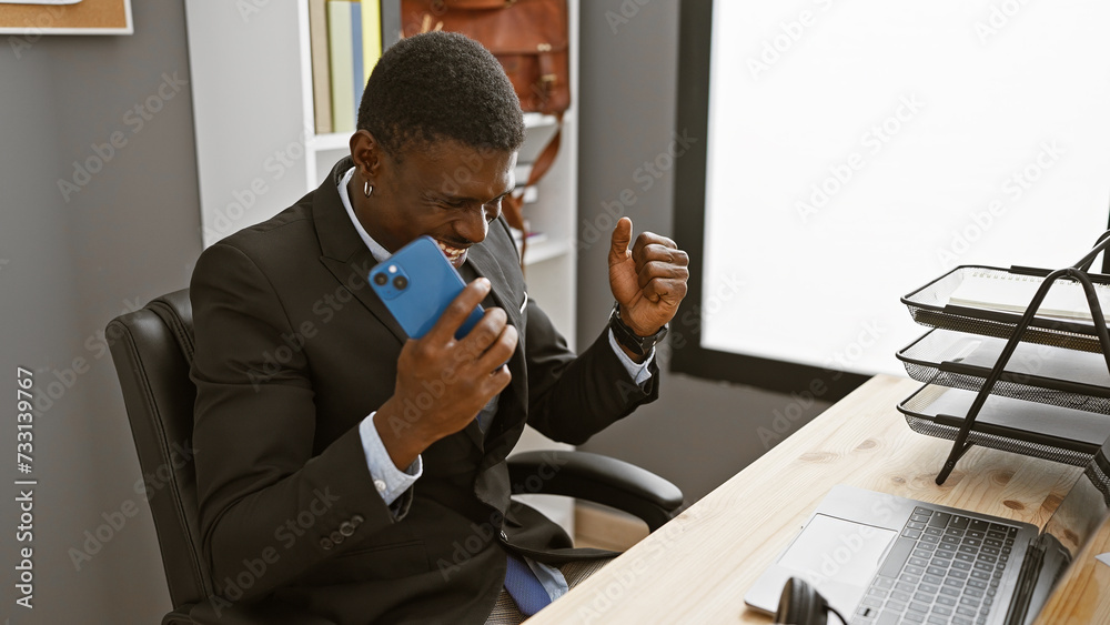 An african american man in a suit celebrates with a smartphone in an office setting, exuding professionalism and joy.