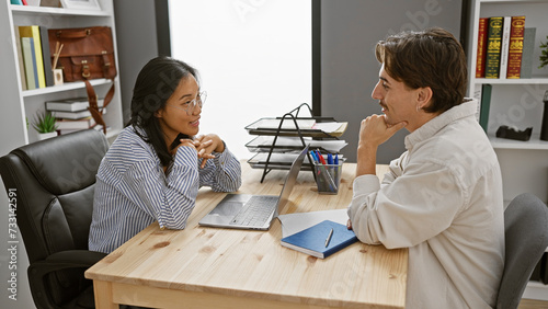 A woman and a man, likely coworkers, engage in conversation at an office with a laptop and notepad on the desk.