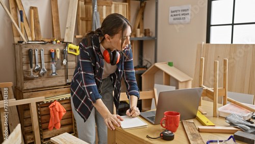 A focused young woman sketches designs in her carpentry workshop surrounded by tools and wood.