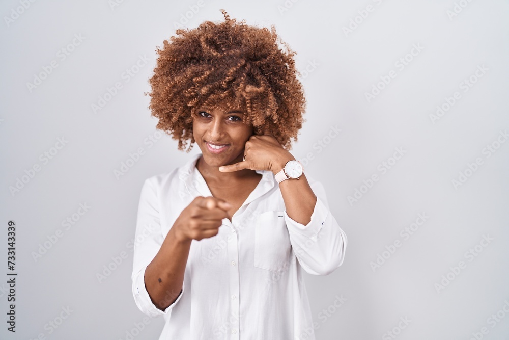 Φωτογραφία Young hispanic woman with curly hair standing over white background smiling doing talking on the telephone gesture and pointing to you