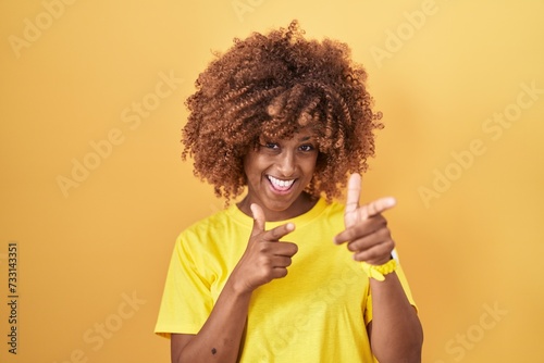 Young hispanic woman with curly hair standing over yellow background pointing fingers to camera with happy and funny face. good energy and vibes.