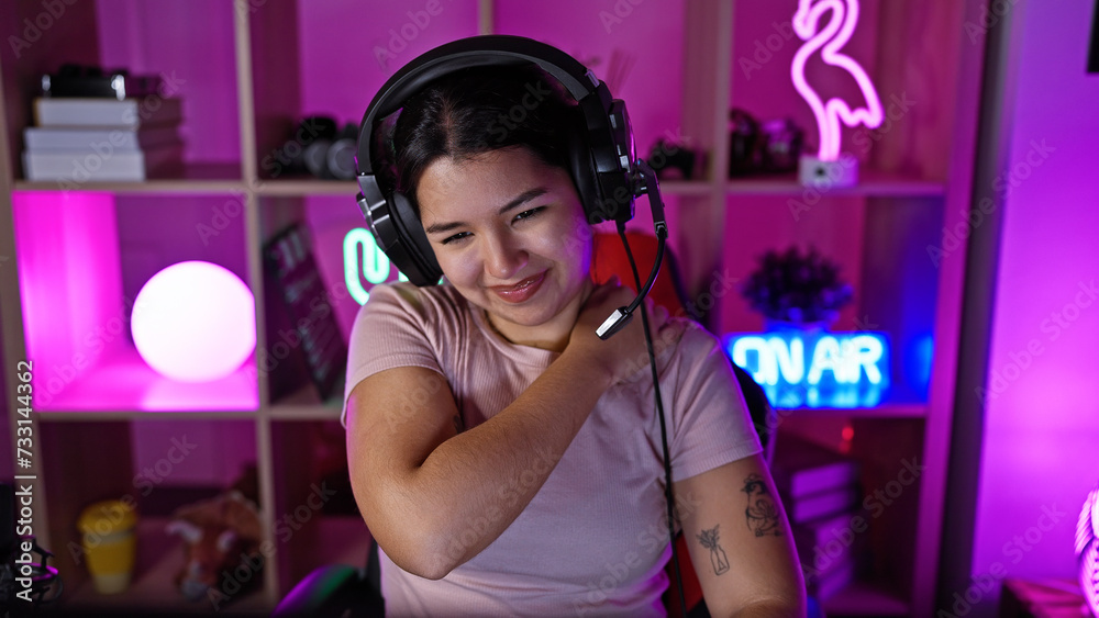 A young hispanic woman with headphones grimaces due to neck pain in a neon-lit gaming room at night.
