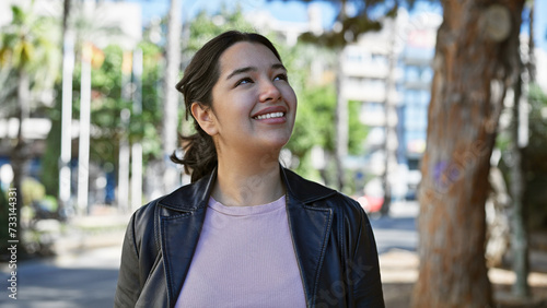 A smiling young hispanic woman, looking away thoughtfully outdoors surrounded by greenery on a city street.