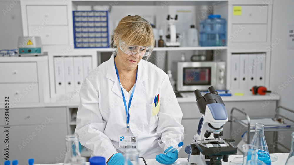 Serious, blonde middle-age woman scientist, taking notes and holding a test tube in a bustling lab, immersed in groundbreaking medical research Serious, blonde middle-age woman scientist, taking notes and holding a test tube in a bustling lab, immersed in groundbreaking medical research