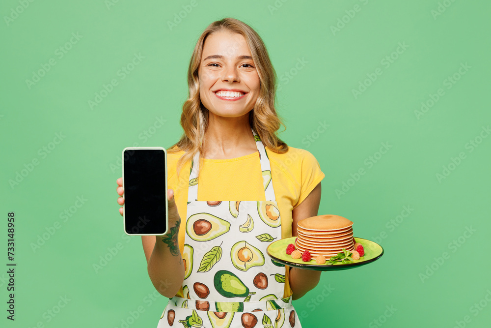 Young housewife housekeeper chef cook baker woman wear apron yellow t-shirt hold plate with pancakes use blank screen mobile cell phone isolated on plain pastel green background. Cooking food concept. Young housewife housekeeper chef cook baker woman wear apron yellow t-shirt hold plate with pancakes use blank screen mobile cell phone isolated on plain pastel green background. Cooking food concept.