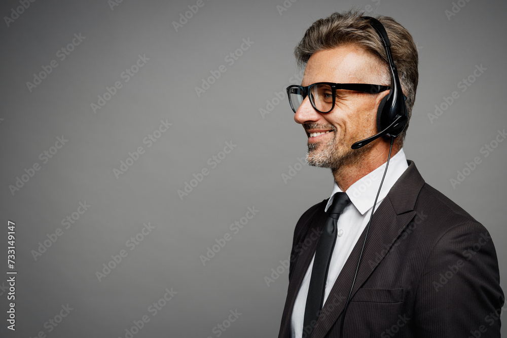 Close up adult employee operator business man wears black suit set microphone headset for helpline assistance sit work at call center office desk with pc computer isolated on grey background studio. Close up adult employee operator business man wears black suit set microphone headset for helpline assistance sit work at call center office desk with pc computer isolated on grey background studio.