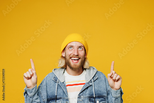 Young smiling happy fun blond man he wear denim shirt hoody beanie hat casual clothes point index finger overhead on area mockup isolated on plain yellow background studio portrait. Lifestyle concept