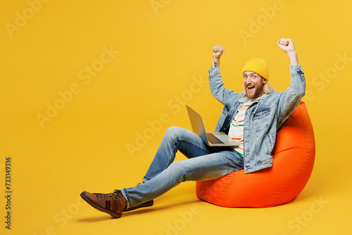 Full body young IT man wears denim shirt hoody beanie hat casual clothes sit in bag chair hold use work on laptop pc computer do winner gesture isolated on plain yellow background. Lifestyle concept. photo