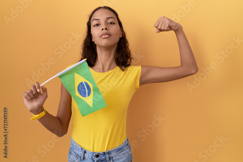 Young hispanic woman holding brazil flag strong person showing arm muscle  confident and proud of power