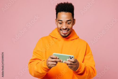 Young man of African American ethnicity wear yellow hoody casual clothes play racing app on mobile cell phone gadget smartphone for pc video games isolated on plain pink background Lifestyle concept.