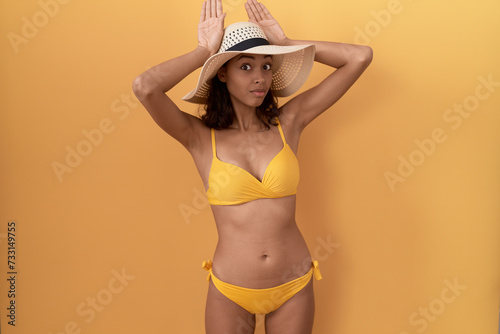 Young hispanic woman wearing bikini and summer hat doing bunny ears gesture with hands palms looking cynical and skeptical. easter rabbit concept. photo