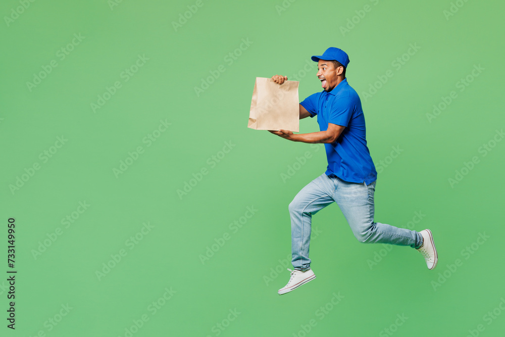 Full body delivery guy employee man in blue cap t-shirt uniform workwear work as dealer courier hold clear blank craft paper takeaway bag jump high isolated on plain green background Service concept