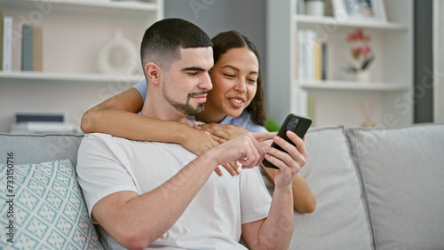 Smiling, confident couple delight in fun phone time, beautiful man and woman, together on their lovely sofa. enjoying the casual lifestyle at home, theyâ€™re texting & hugging, indoors