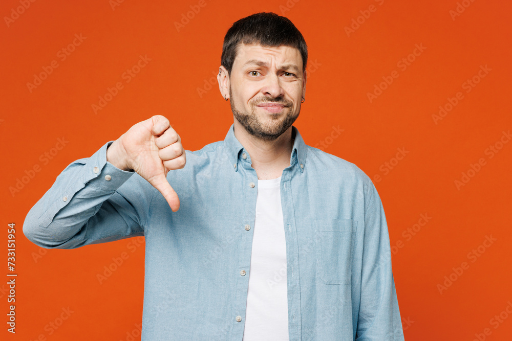 Young dissatisfied displeased sad man he wearing blue shirt white t-shirt casual clothes showing thumb down dislike gesture isolated on plain red orange background studio portrait. Lifestyle concept. Young dissatisfied displeased sad man he wearing blue shirt white t-shirt casual clothes showing thumb down dislike gesture isolated on plain red orange background studio portrait. Lifestyle concept.