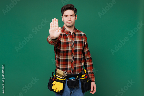 Young employee laborer handyman man wear red shirt showing stop hand gesture with palm isolated on plain green background. Instruments accessories for renovation apartment room. Repair home concept.